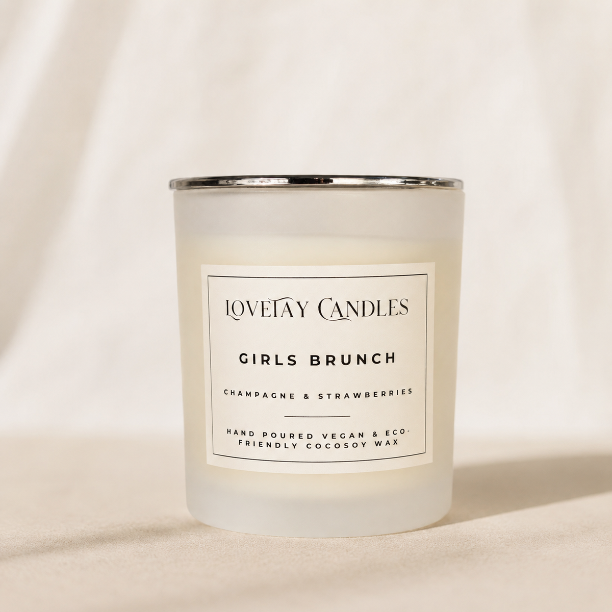 A frosted glass candle jar with a silver metal rim and a white label reading 'LOVETAY CANDLES GIRLS BRUNCH' on a warm beige fabric background.