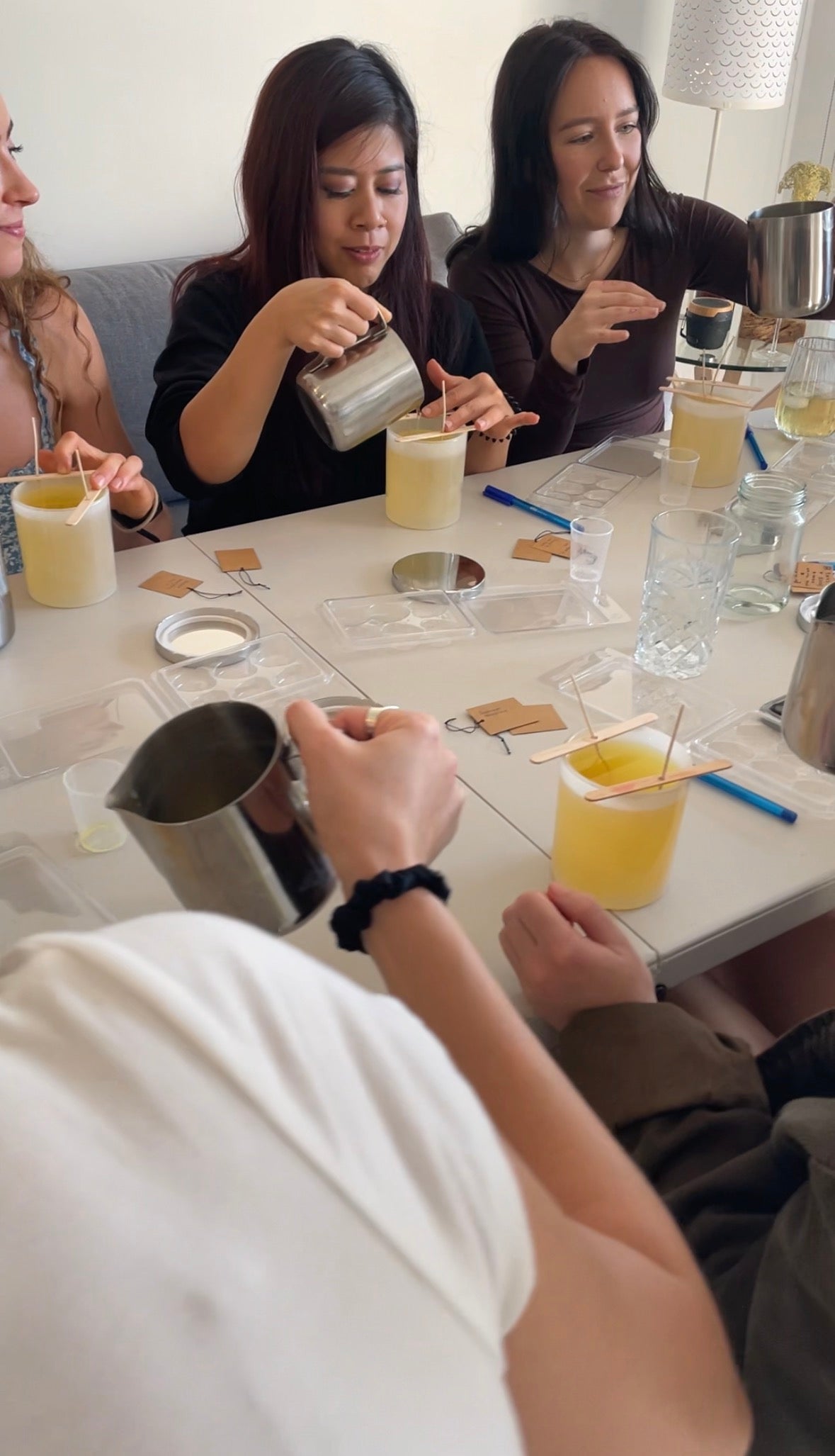 Three women sitting around a table with drinks and snacks, one of them is pouring wax to make her own LoveTay Candle. .