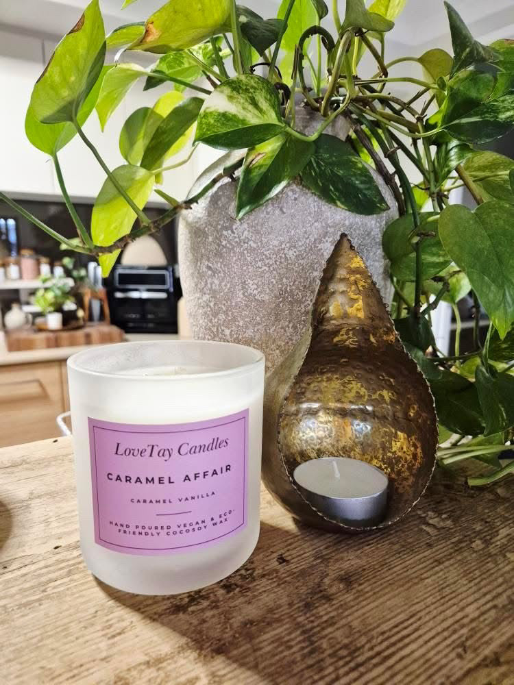 Candle labeled 'Caramel Affair' by LoveLay Candles on a wooden surface with plants in the background.