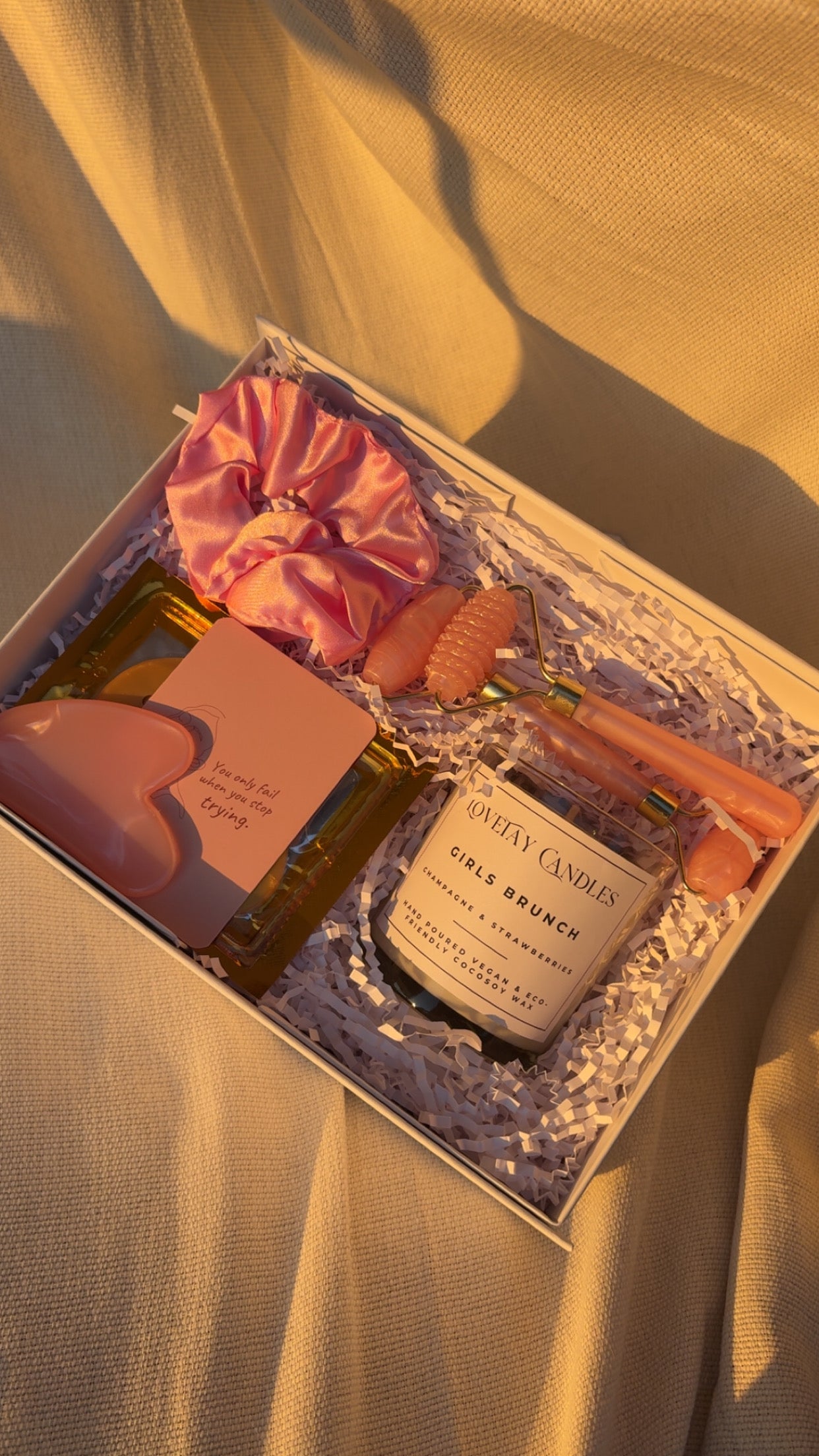 Gift set with a candle, eye mask, face roller set, affirmation card, and a scrunchie in a decorative box on a soft fabric background.