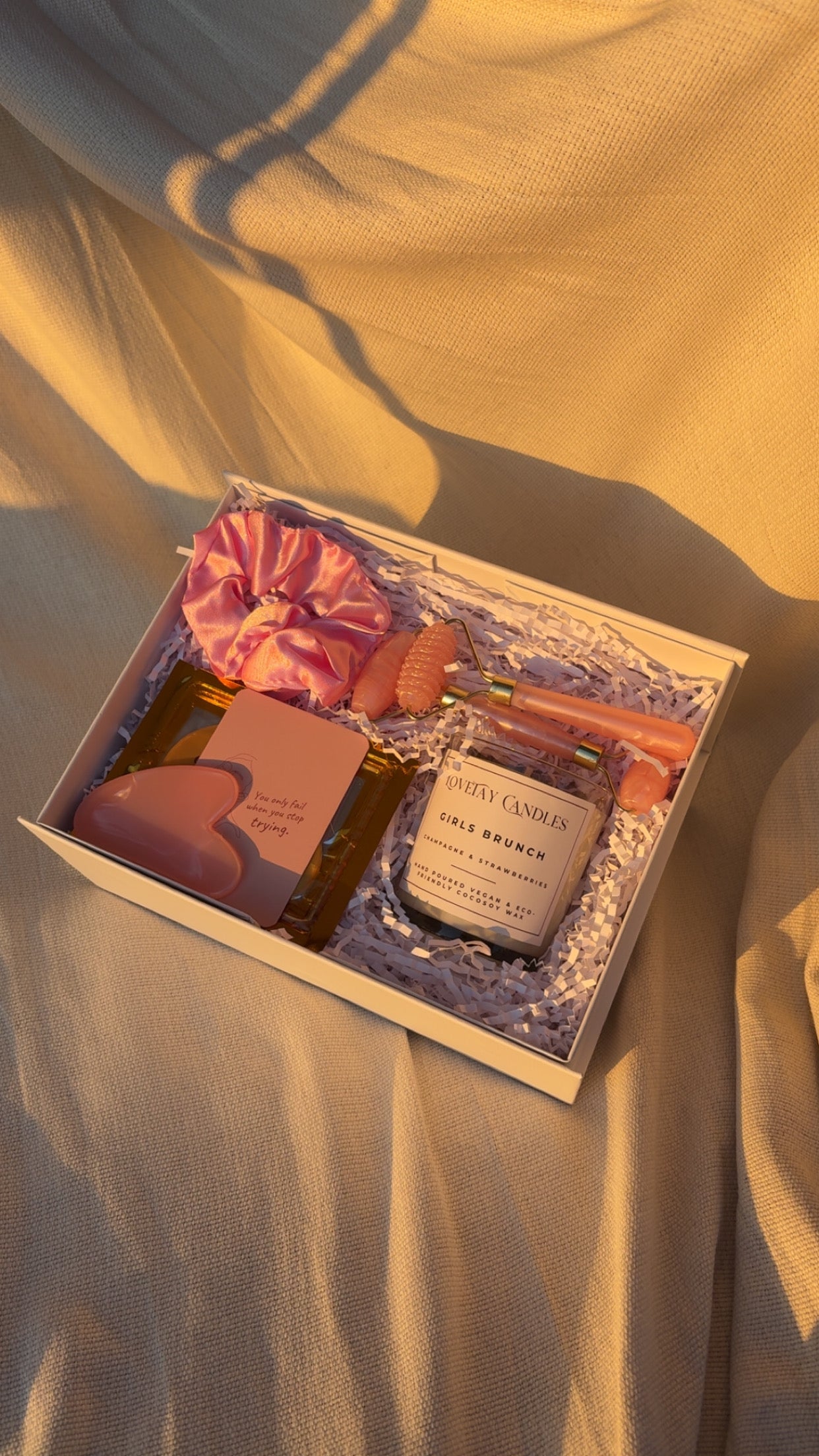 Gift set with a candle, eye mask, face roller set, affirmation card, and a scrunchie in a decorative box on a soft fabric background.
