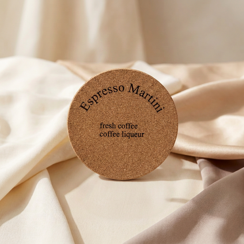 A round cork coaster with the words 'Espresso Martini' and 'fresh coffee coffee liqueur' printed on it, resting on draped cream-colored fabric.