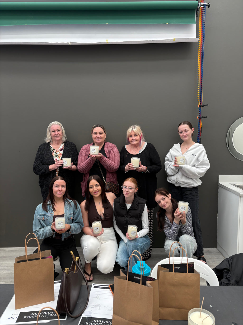 A group of nine women posing together in an indoor LoveTay Candles workshop space, each holding a small white LoveTay candle. Several brown paper shopping bags and printed cards are placed on a table in the foreground against a dark gray wall.