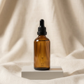 An amber glass dropper bottle with a black rubber bulb and black ribbed cap, standing on a small white pedestal against a draped beige fabric background.