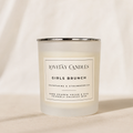 A frosted glass candle jar with a silver metal rim and a white label reading 'LOVETAY CANDLES GIRLS BRUNCH' on a warm beige fabric background.