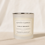 A frosted glass candle jar with a silver metal rim and a white label reading 'LOVETAY CANDLES GIRLS BRUNCH' on a warm beige fabric background.