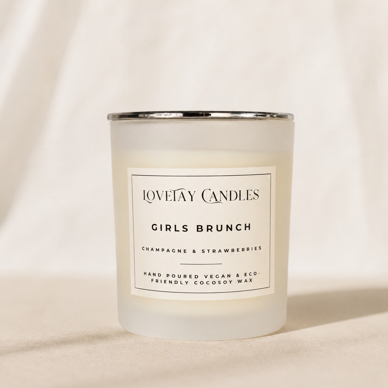 A frosted glass candle jar with a silver metal rim and a white label reading 'LOVETAY CANDLES GIRLS BRUNCH' on a warm beige fabric background.