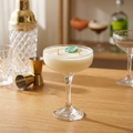 A creamy off-white cocktail in a coupe glass, garnished with a small green leaf, set on a wooden bar table with a shaker, jigger, and other bar tools blurred in the background.