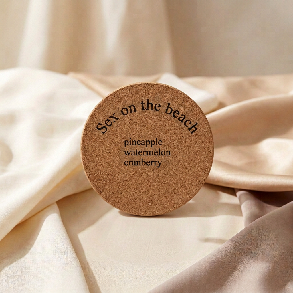 A round cork coaster with the words 'Sex on the beach' and flavor names 'pineapple', 'watermelon', and 'cranberry' printed on it, resting on draped cream-colored fabric.