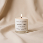 A lit glass jar candle labeled 'LOVETAY CANDLES SUMMER TIME Juicy Watermelon' sits on draped beige fabric. The candle has a white label with black text and is shown in a warm, softly lit setting.