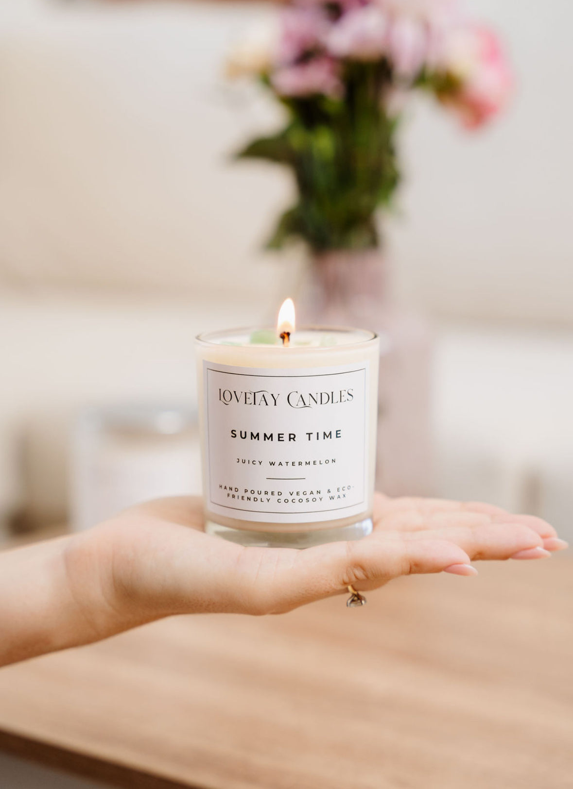 Taylah's hand holding a lit candle labelled 'Summer Time' by LoveTay Candles with a blurred background of flowers and furniture.
