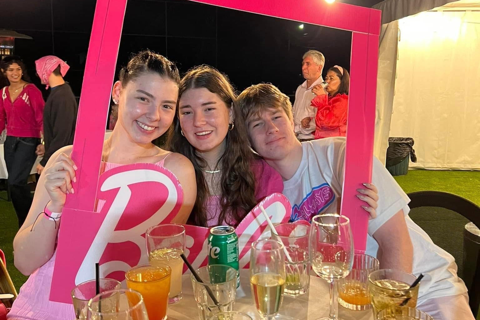People posing with a pink frame at a social gathering with drinks on a table.