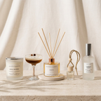 A styled collection of LoveTay Candles home fragrance products on a stone surface against a draped beige fabric backdrop. The set includes a Girls Brunch scented candle, a reed diffuser with a wooden reed stick and rose gold cap, a small reed diffuser bottle with a jute loop, and a Garden Party room spray with a silver cap.