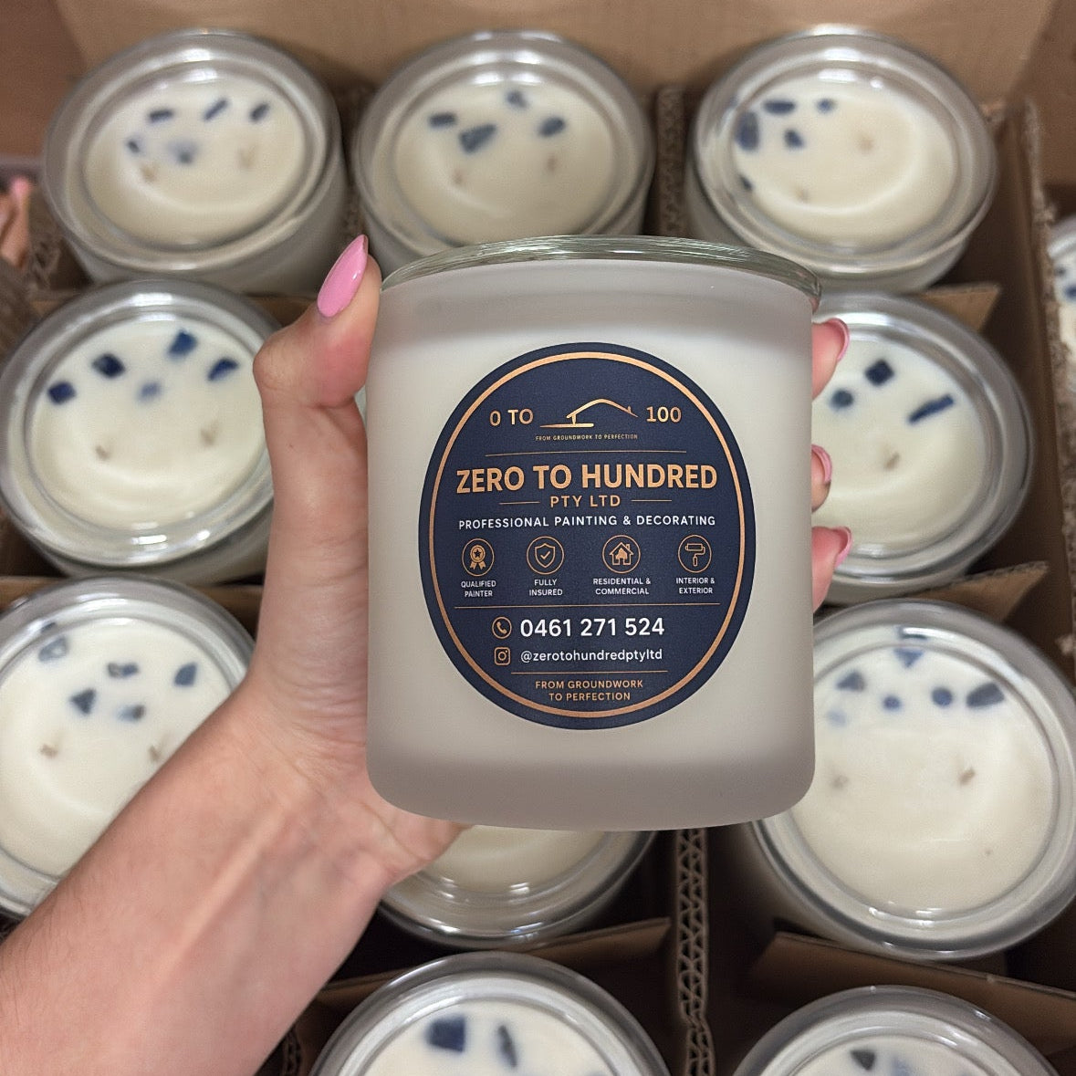 A hand holds a frosted glass jar candle labeled 'Zero to Hundred' in front of a cardboard box filled with multiple identical candles. The candle wax is white with small blue botanical pieces floating on top.
